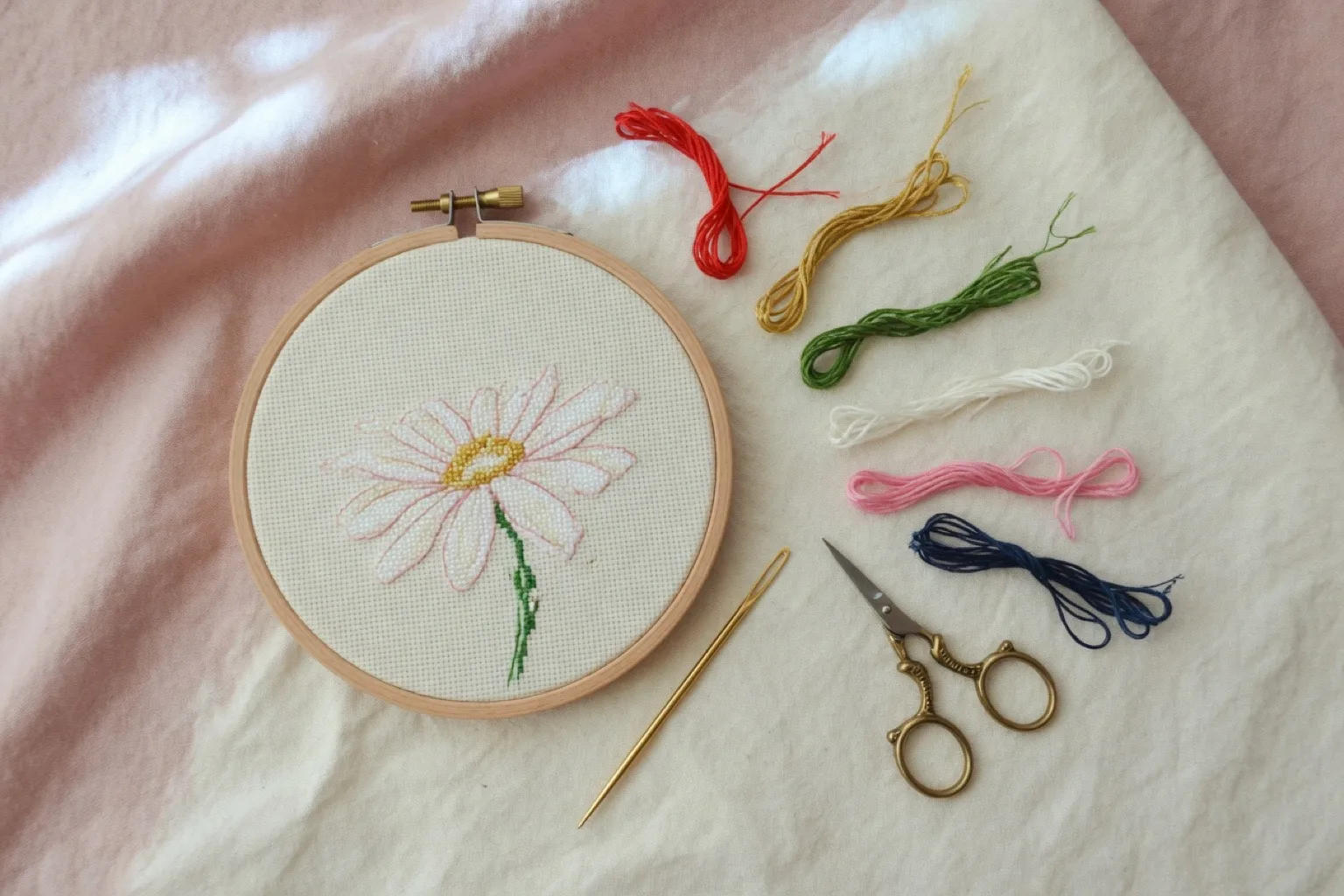 Small hoop with a simple daisy, six colorful floss lengths, a gold needle, and stork scissors on cream cloth