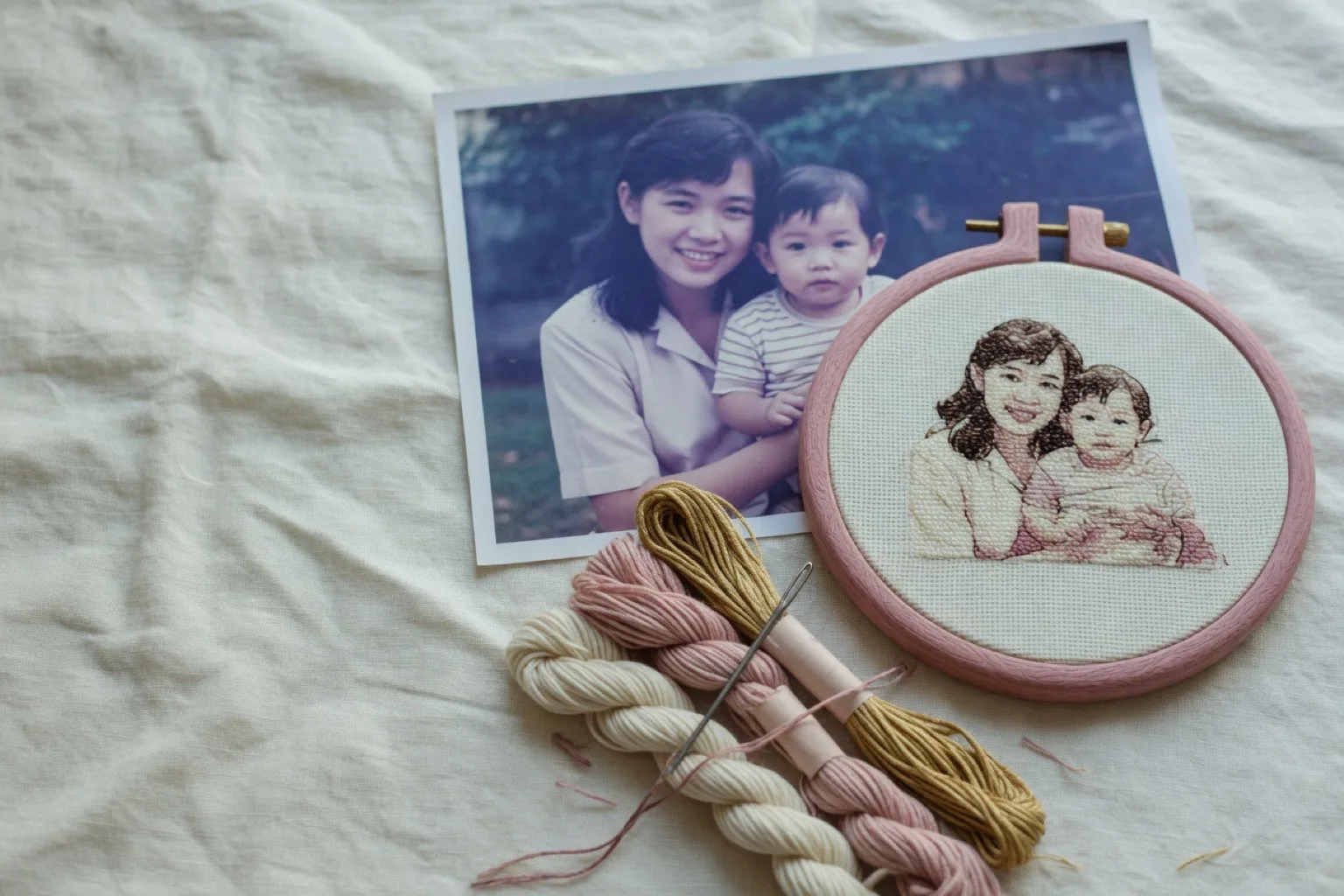 Printed photo of a mother and toddler beside a wooden hoop with the same pair stitched in cream, brown, and rose thread, with matching floss skeins on linen